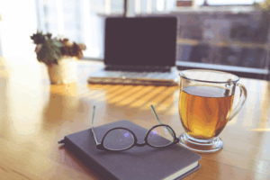 A laptop computer, a mug of tea, a notebook and a pair of glasses sitting on a desk.
