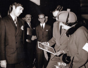 LifeBook author Derek Rose at the British Menswear Guild exhibition, 1966, showing Queen Elizabeth II a photograph of a pair of pajamas selected by King George VI at the British Industries Fair, 1937. 